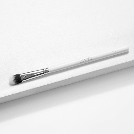 Mini Duo Fiber Face Brush by ColourPop resting diagonally on a white textured surface, with a glossy white handle, silver ferrule, and soft black-to-white duo-fiber bristles for blending and buffing face makeup.