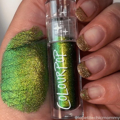 ColourPop Chrome Liquid Shadow in Beetle shows a bold emerald-to-gold shift, swatched across the back of a hand as gold-glitter tipped nails hold the clear tube with starry cap, showcasing the high-shine, multidimensional eye shadow finish.