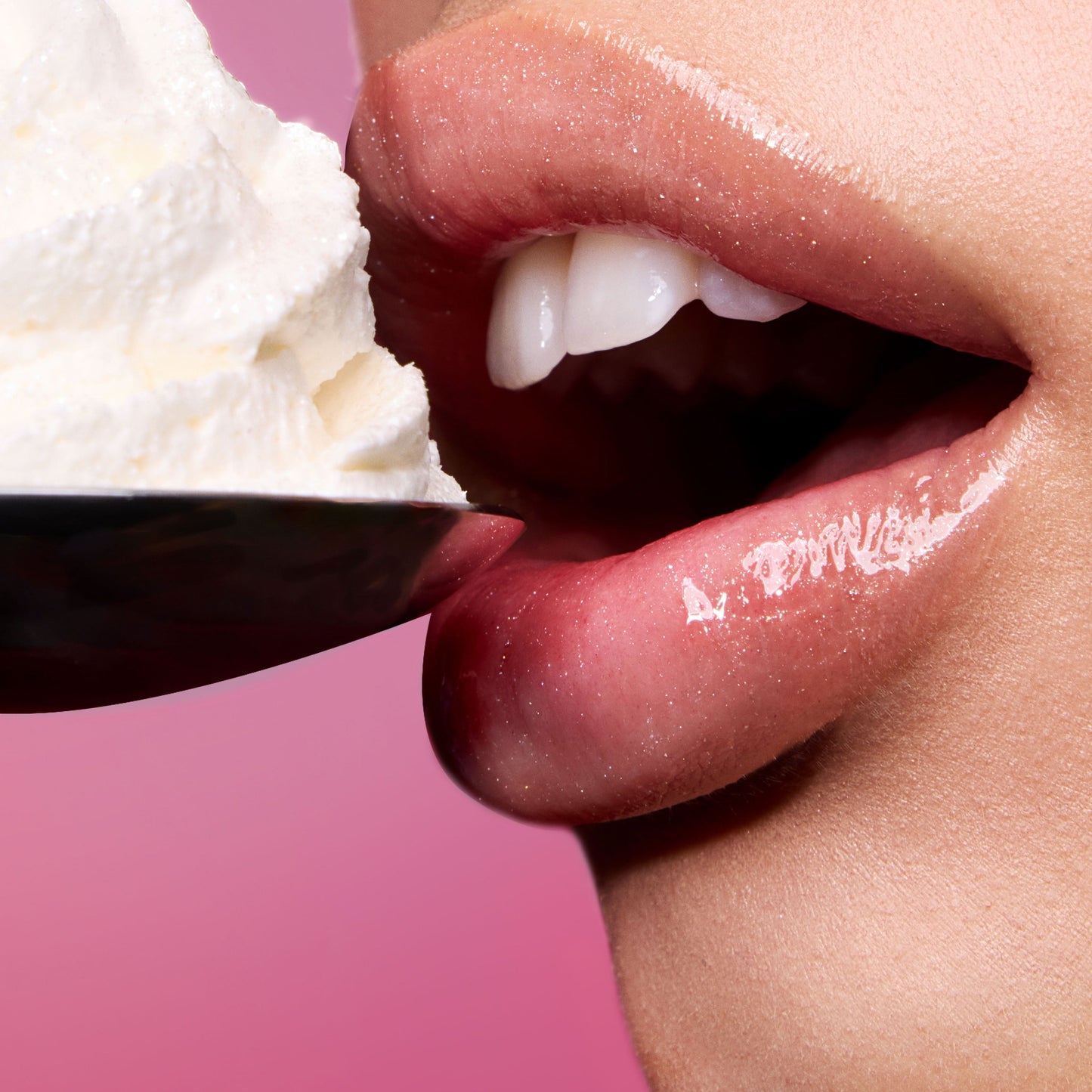 Close-up glossy lips wearing Colourpop Fresh Kiss Glossy Balm in Whipped Cream hover near a spoonful of whipped cream against a pink background, showcasing the shimmery Lip Gloss & Lip Oil texture with slogan “You look good enough to eat.”