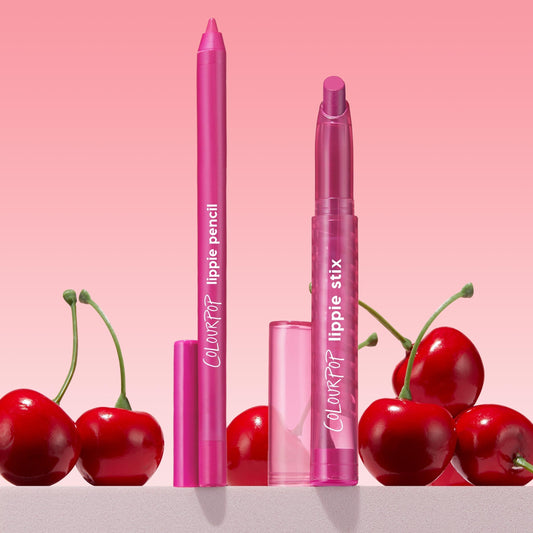Heart Lips ColourPop Lippie Stix + Pencil Set stands upright with caps off, revealing hot-pink lipstick and matching liner in translucent pink tubes labeled ColourPop, styled among glossy red cherries on a pink gradient background.