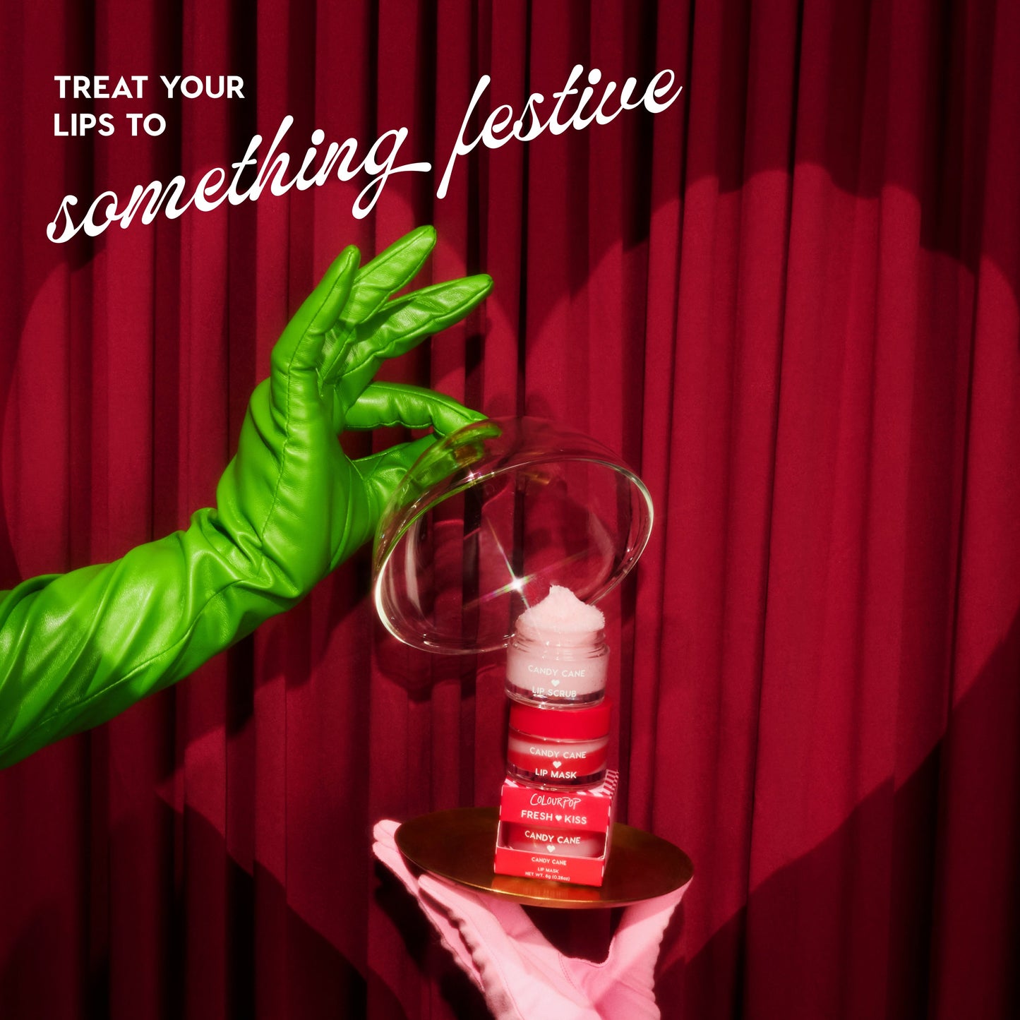 Green-gloved hand holding a lip balm against a red curtain with text 'Treat your lips to something festive'.