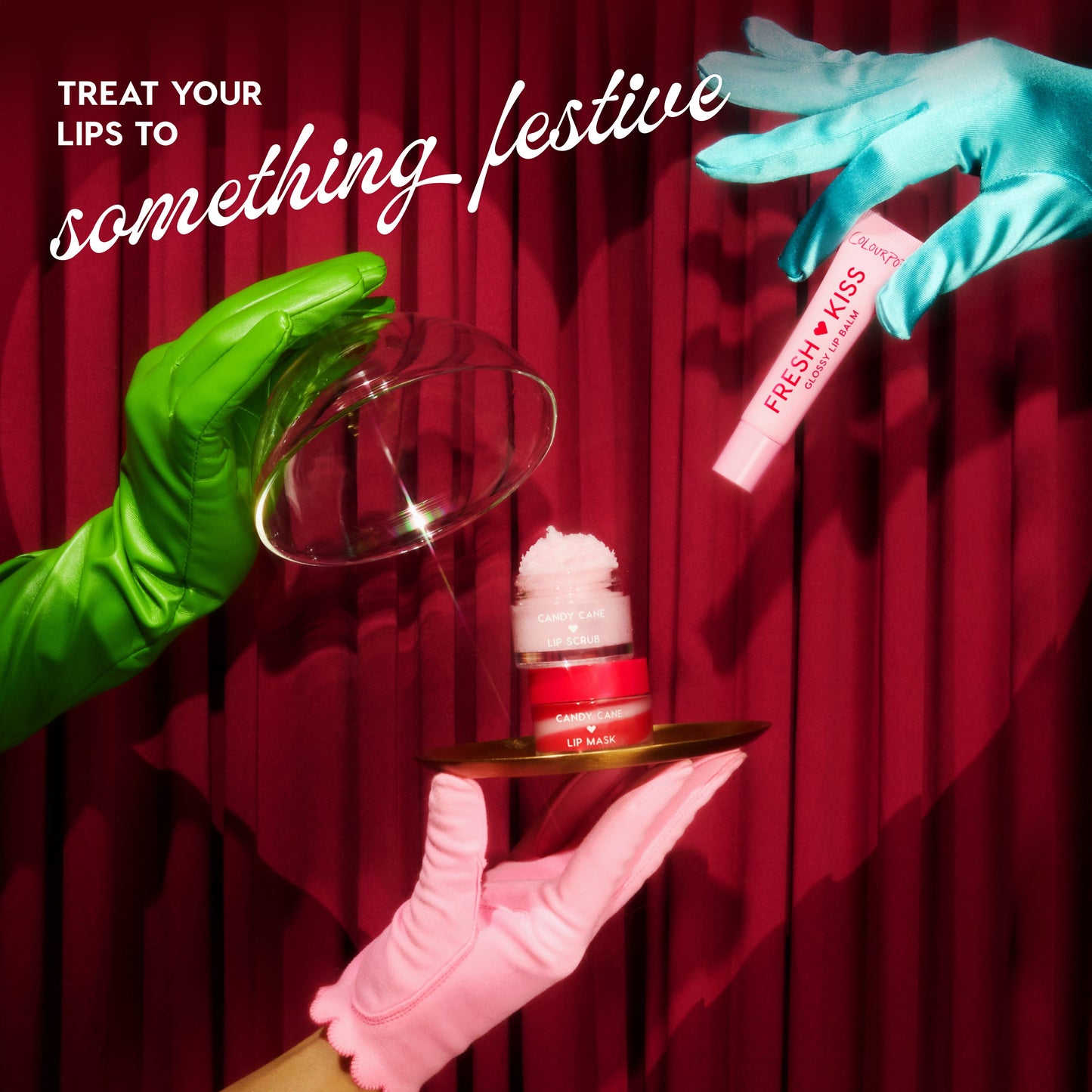 Mistletoe Kisses lip care kit by Colourpop - Fresh Kiss glossy lip balm and Candy Cane Lip Scrub + Lip Mask - presented by green, blue, and pink gloved hands lifting a glass cloche over a gold tray against a red holiday curtain.