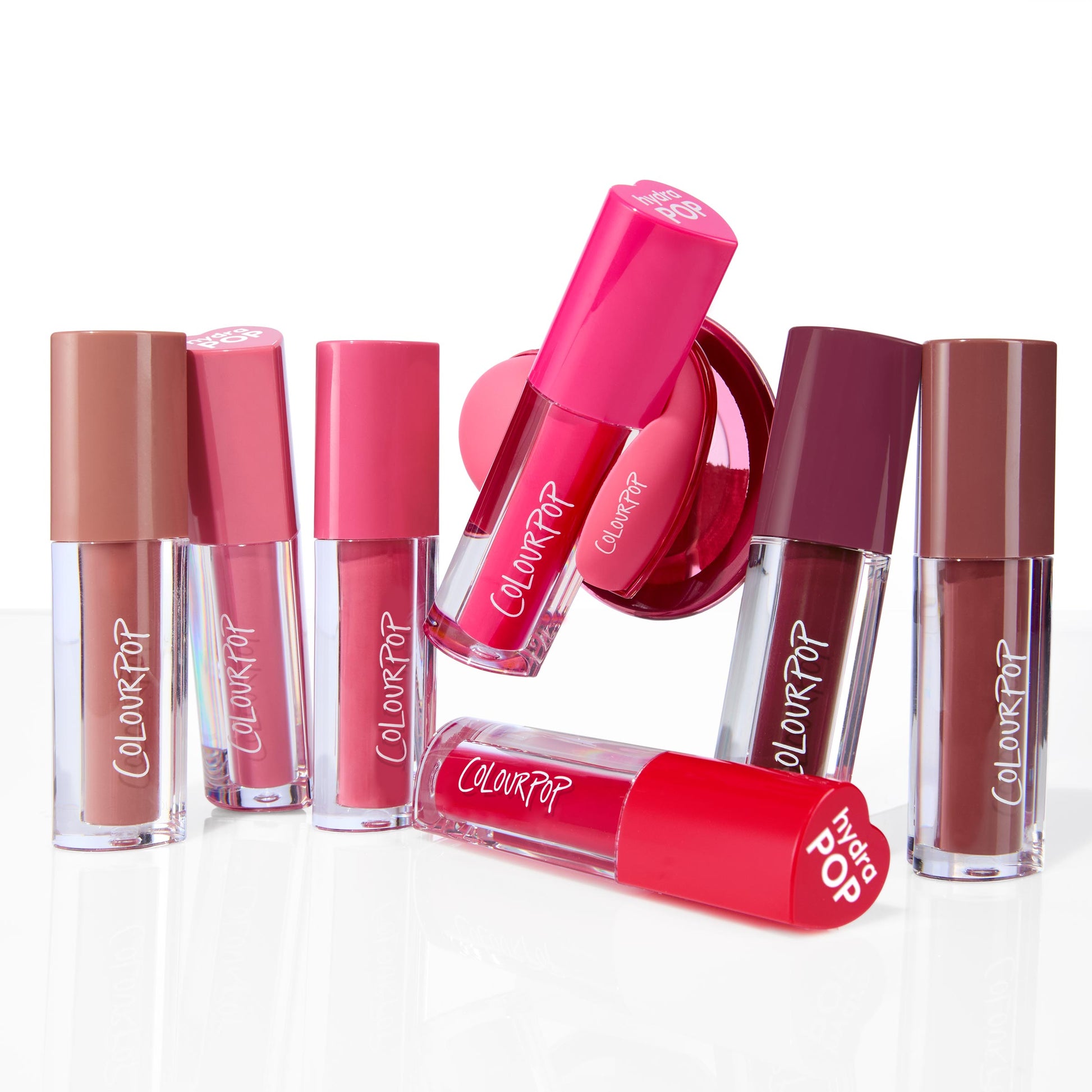 ColourPop Lip Stain Pop Stars tubes standing, leaning, and one lying to show nude, pink, and berry shades, paired with a hot‑pink Hydra Pop PopSocket phone grip, arranged on a glossy white studio surface.