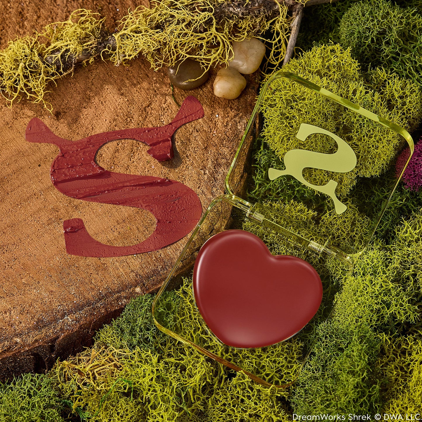Fairytale Things ColourPop Lip and Cheek Balm sits open, revealing a heart-shaped pan of warm brick-red pigment in a translucent green, Shrek-inspired compact with an ogre-ear S, resting on moss and a wood slice beside a glossy burgundy S swatch.