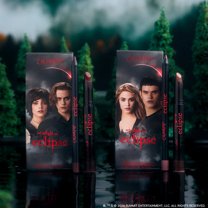 ColourPop Twilight Immortal Kiss Lippie Stix Duo stands upright on a reflective lake surface, with Eclipse-themed character boxes and matching deep nude and berry lip pencils and sticks framed by a dark, misty forest backdrop.