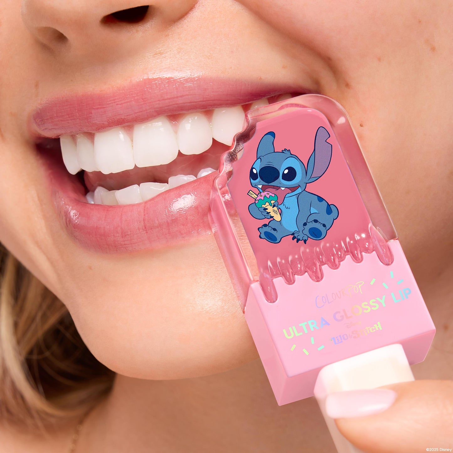 ColourPop Ultra Glossy Lip in Super Sweet coats a smiling mouth with sheer pink shine, the pink tube with Disney Stitch art held to glossy lips in a tight close-up, showcasing the high-shine finish and playful packaging.