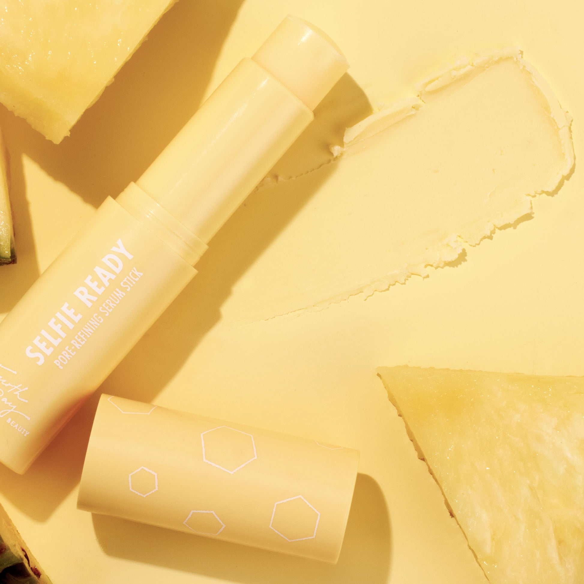 Selfie Ready pore-refining ColourPop Serum Stick resting open with its cap beside it and a creamy swipe, on a monochromatic yellow backdrop styled with pineapple slices, showcasing the portable serum stick’s smooth texture and bright vibe.